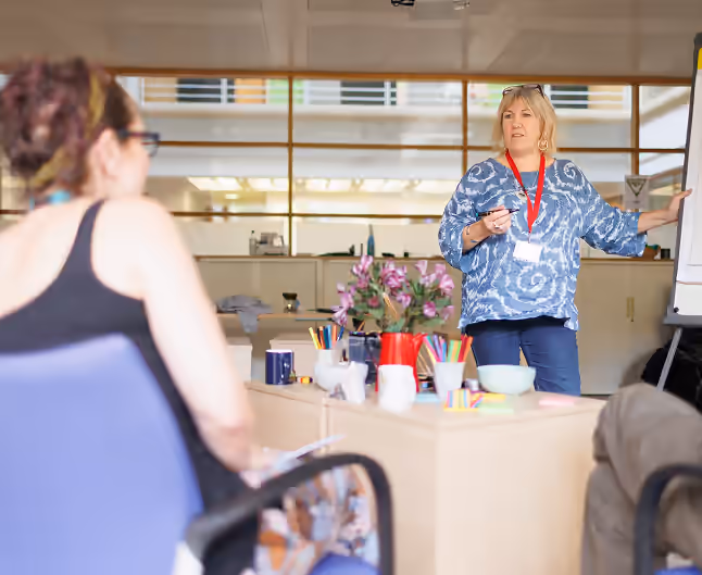 A female trainer in a blue patterned shirt presents to a class, pointing to a whiteboard during a CPD-accredited communication course.