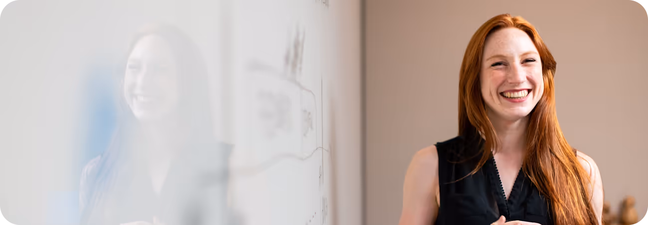 A cheerful woman with long red hair smiles warmly at the camera while standing next to a whiteboard in an office setting, representing NFS Mediation’s Coaching Plus Service.