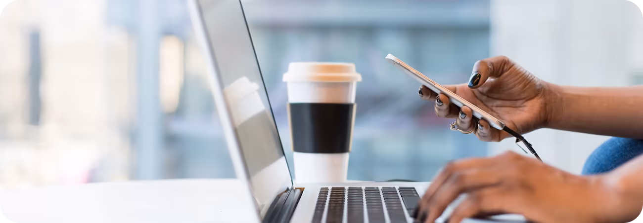 Close-up of a person's hands using a smartphone and a laptop simultaneously, with a coffee cup on the desk, representing contacting NFS Mediation.